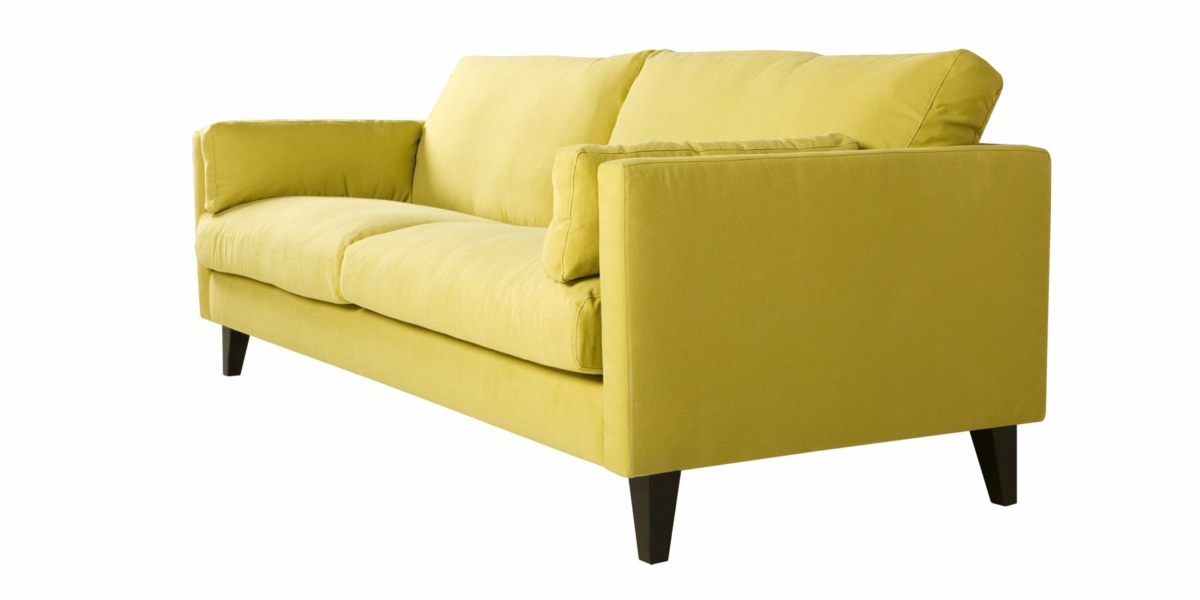 Un canapé jaune chic avec des coussins moelleux et des pieds en bois foncé se dresse élégamment sur un fond blanc, dégageant un charme parisien. Canape Paris by Atelier Bis