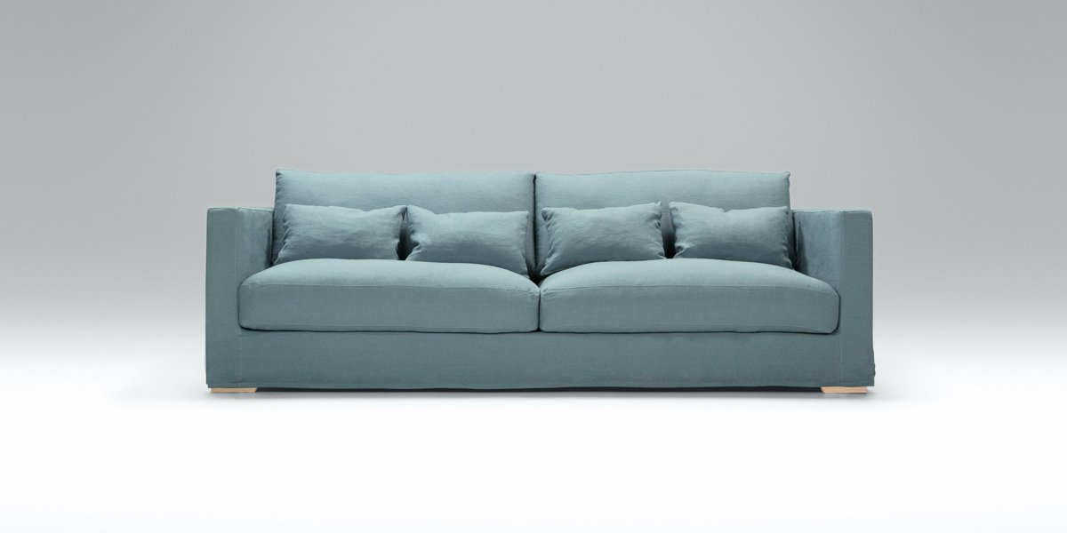 Canapé moderne bleu clair avec deux grands coussins et deux petits oreillers posés sur un fond gris uni, rappelant les intérieurs chics de Paris. Canape Paris by Atelier Bis