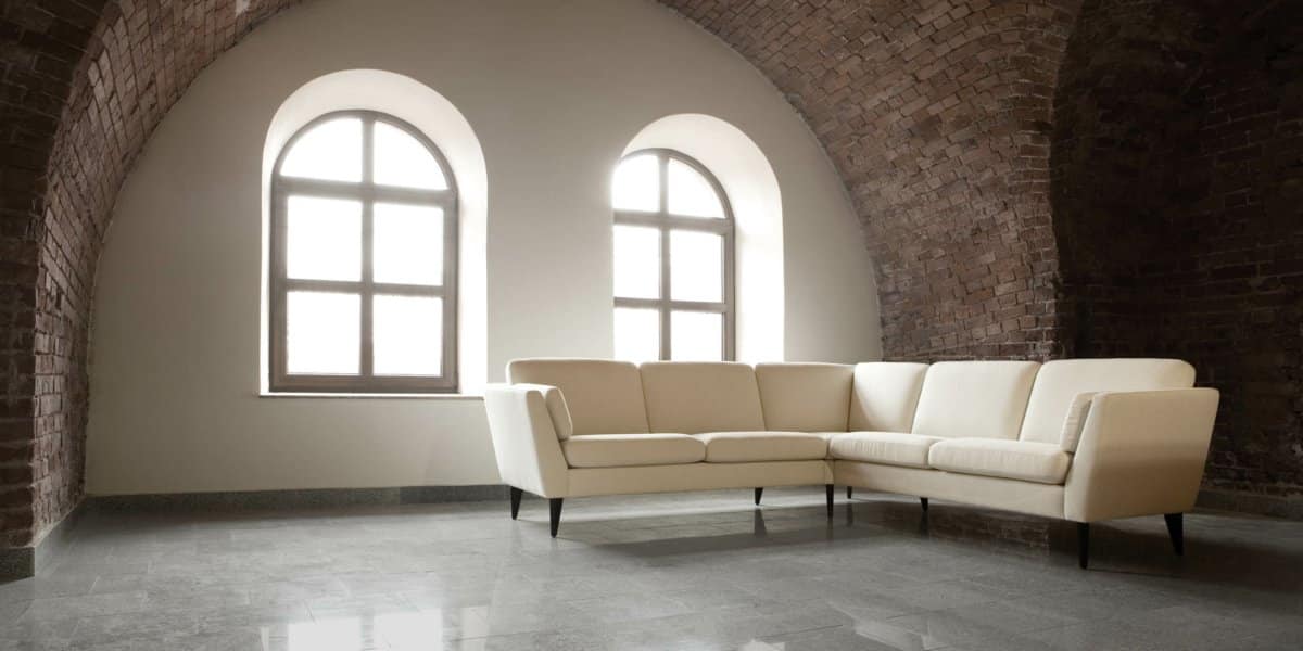 Chambre au plafond voûté en briques avec deux grandes fenêtres rappelant le charme parisien, avec un canapé d'angle beige sur un sol gris brillant. Canape Paris by Atelier Bis