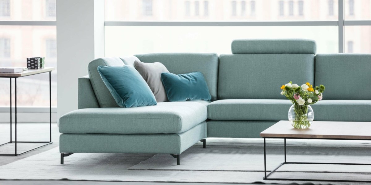 Un salon chic et moderne présente un canapé vert clair accentué par des coussins bleu sarcelle et gris, complété par une table basse élégante ornée d'un vase à fleurs, évoquant l'élégance d'une retraite parisienne. Canape Paris by Atelier Bis