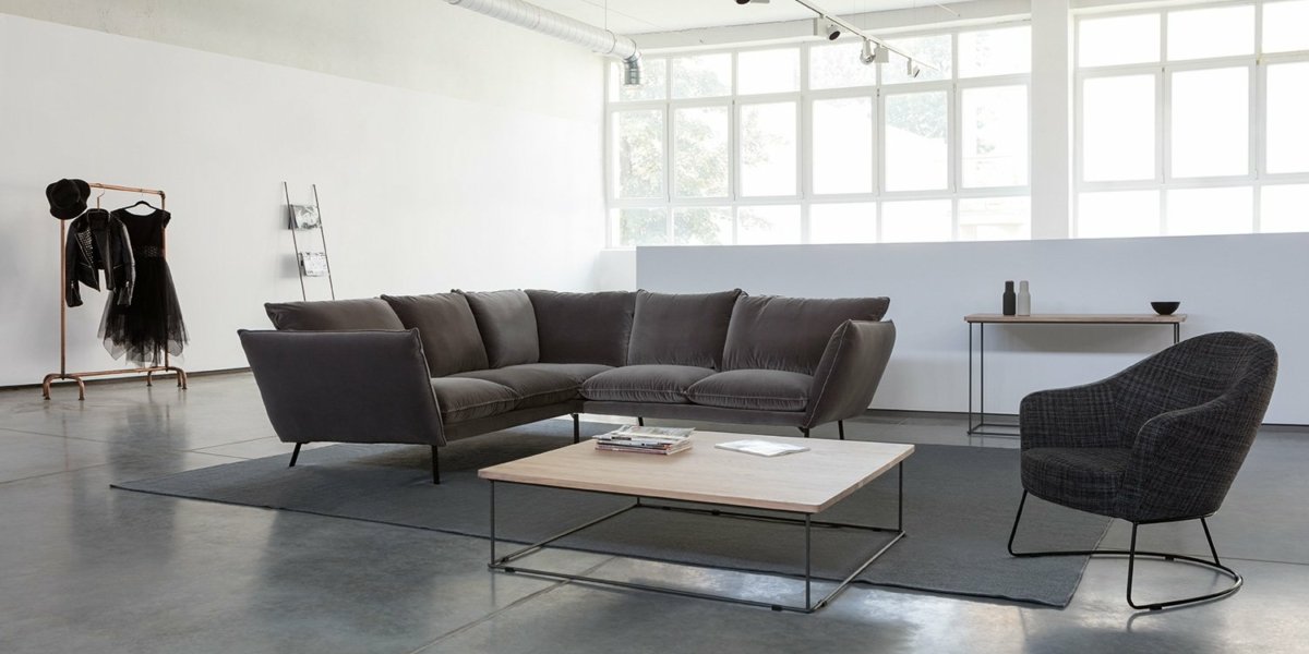 Un salon minimaliste d'inspiration parisienne comprend un canapé d'angle gris, un fauteuil et une table basse sur un grand tapis, avec un portant gracieusement niché à l'arrière. Canape Paris by Atelier Bis