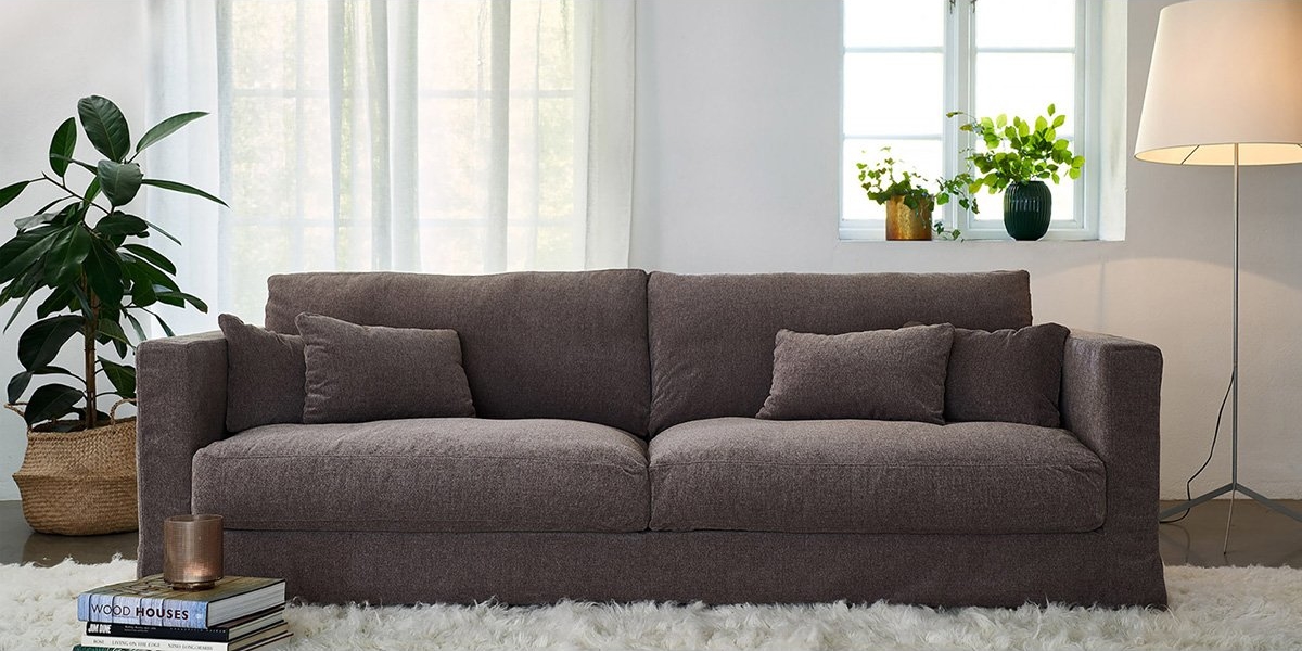 Un canapé gris de qualité avec des oreillers repose sur un tapis moelleux, entouré de plantes, de livres et d'une lampe près de deux fenêtres avec des rideaux lumineux. Canape Paris by Atelier Bis