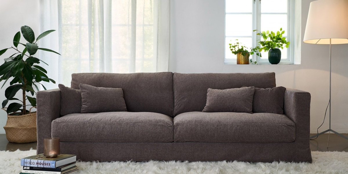 Un canapé marron dans une pièce lumineuse avec un tapis à poils longs, des plantes, une pile de livres et une lampe sur pied à côté d'une fenêtre. Canape Paris by Atelier Bis