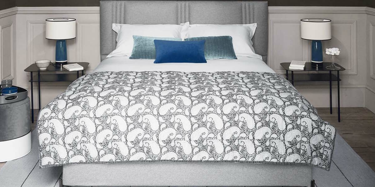 Chambre moderne avec un lit gris, une literie à motifs et des coussins bleus, rappelant un showroom parisien. Deux tables de nuit et des lampes assorties complètent l'ambiance chic. Canape Paris by Atelier Bis