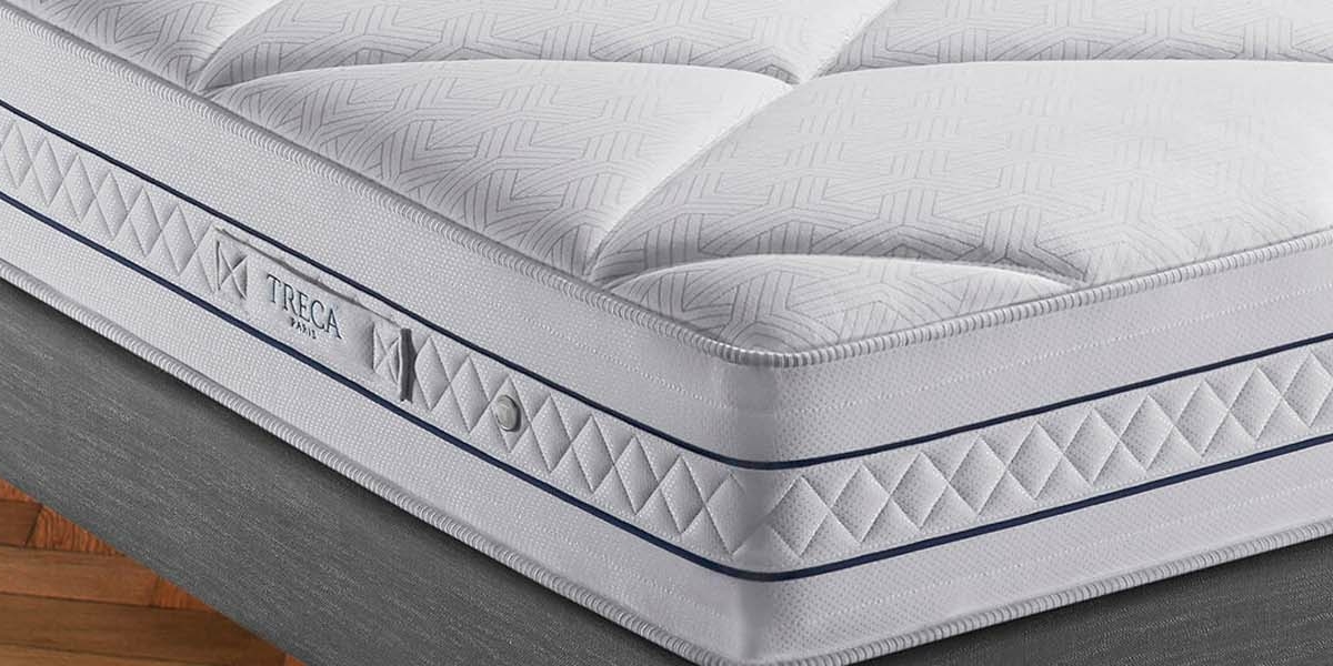 Gros plan d'un matelas soigneusement confectionné avec un tissu blanc à motifs et une bordure bleue sur un plancher en bois, rappelant l'élégance d'une chambre parisienne chic. Canape Paris by Atelier Bis