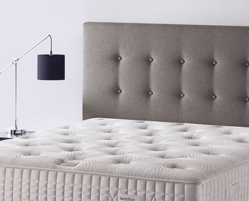 Une tête de lit capitonnée grise avec un design à boutons complète élégamment la lampe moderne et le matelas matelassé blanc, offrant une touche de style inspirée de Paris. Canape Paris by Atelier Bis
