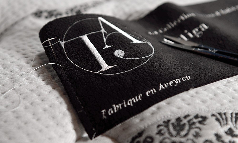 Gros plan d'une étiquette en cuir avec broderie blanche sur une surface textile, rappelant l'élégance parisienne, comportant des ciseaux et des motifs décoratifs. Canape Paris by Atelier Bis