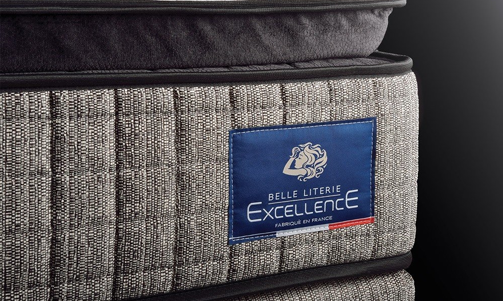 Gros plan d'un matelas gris avec une étiquette bleue indiquant « Belle Literie Excellence, Fabriqué en France », mettant en valeur sa qualité sur un fond sombre. Canape Paris by Atelier Bis