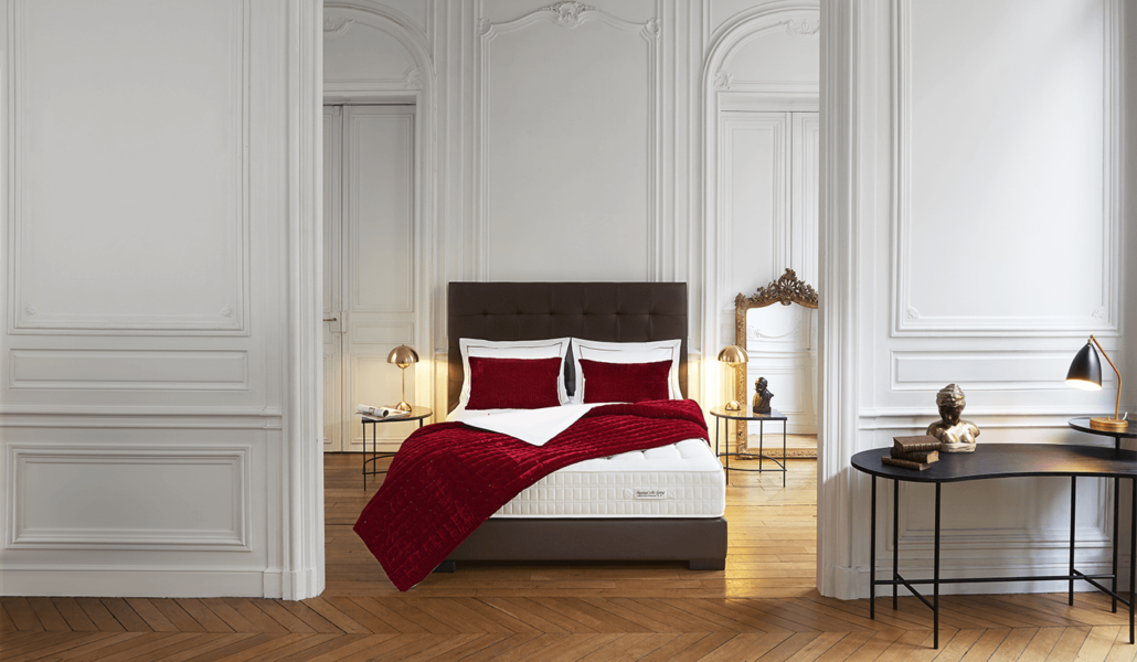 Une chambre parisienne élégante avec un lit doté d'une literie rouge, d'élégants murs blancs, d'un parquet et de lampes modernes illuminant gracieusement le décor chic. Canape Paris by Atelier Bis