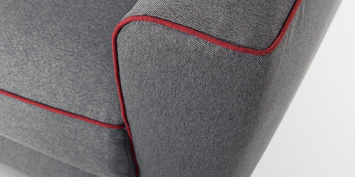 Gros plan d'un accoudoir de canapé en tissu gris avec détail de passepoil rouge, évoquant le chic parisien, sur fond blanc. Canape Paris by Atelier Bis