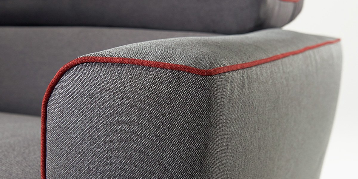 Gros plan d'un accoudoir de canapé en tissu gris avec passepoil rouge sur les bords, mettant en valeur la texture et les détails de conception qui font écho à la qualité parisienne. Canape Paris by Atelier Bis