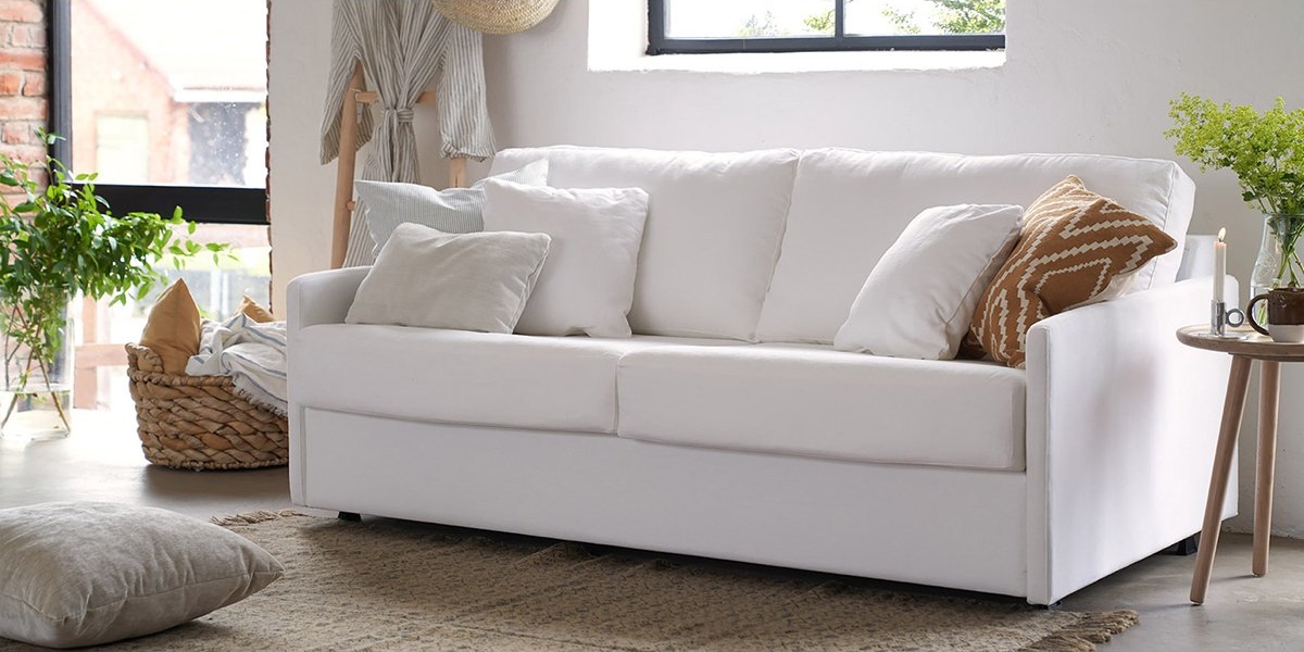 Un salon cosy doté d'un canapé blanc incarnant la qualité parisienne, orné d'oreillers assortis et d'un panier près de la fenêtre mettant en valeur une verdure luxuriante à l'extérieur. Canape Paris by Atelier Bis