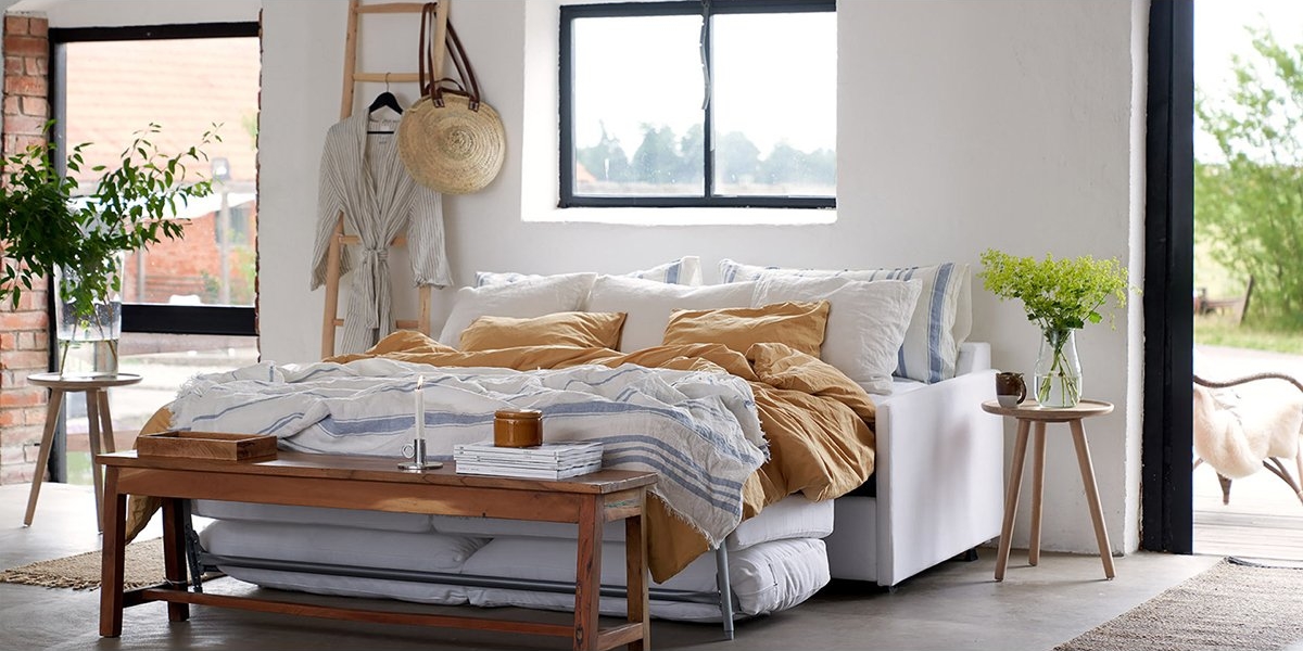 Chambre lumineuse présentant une décoration de qualité salle d'exposition, comprenant un lit avec literie moutarde et blanche, un banc en bois parfait comme canapé, des tables d'appoint ornées de plantes et une vue captivante sur la fenêtre. Canape Paris by Atelier Bis