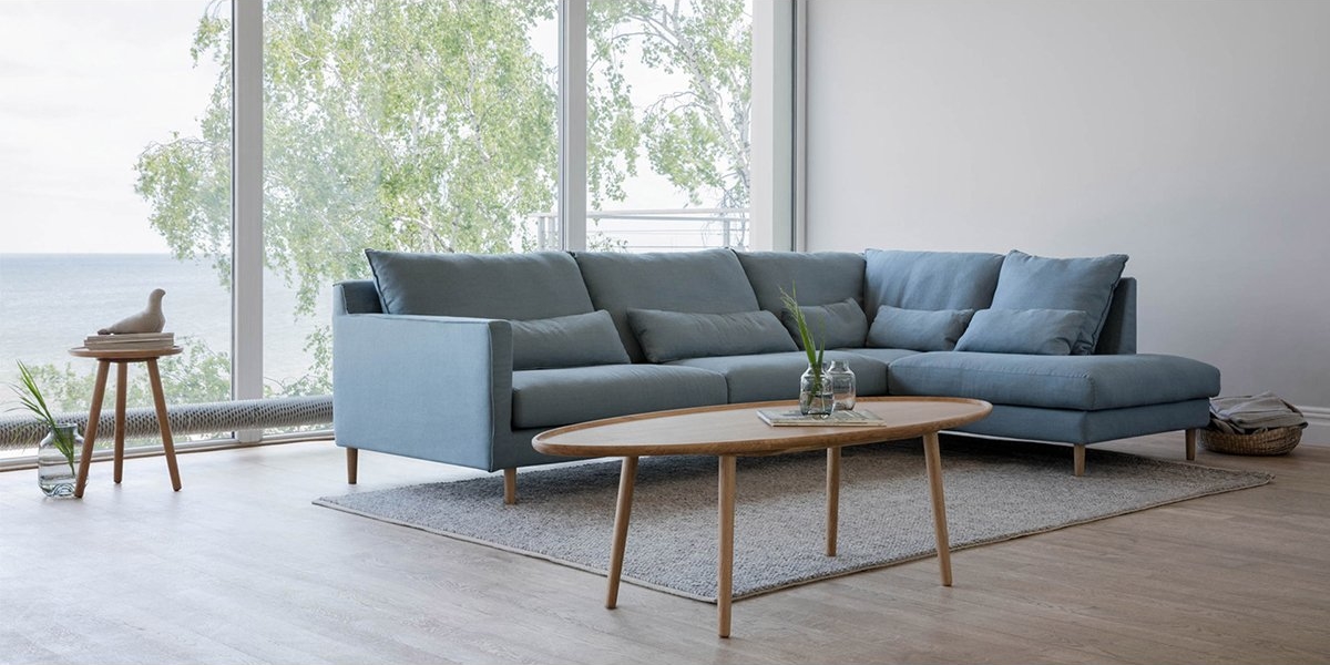 Salon spacieux avec un canapé sectionnel bleu, une table basse en bois et de grandes fenêtres donnant sur la verdure et l'eau - une véritable salle d'exposition parisienne d'élégance et de qualité. Canape Paris by Atelier Bis