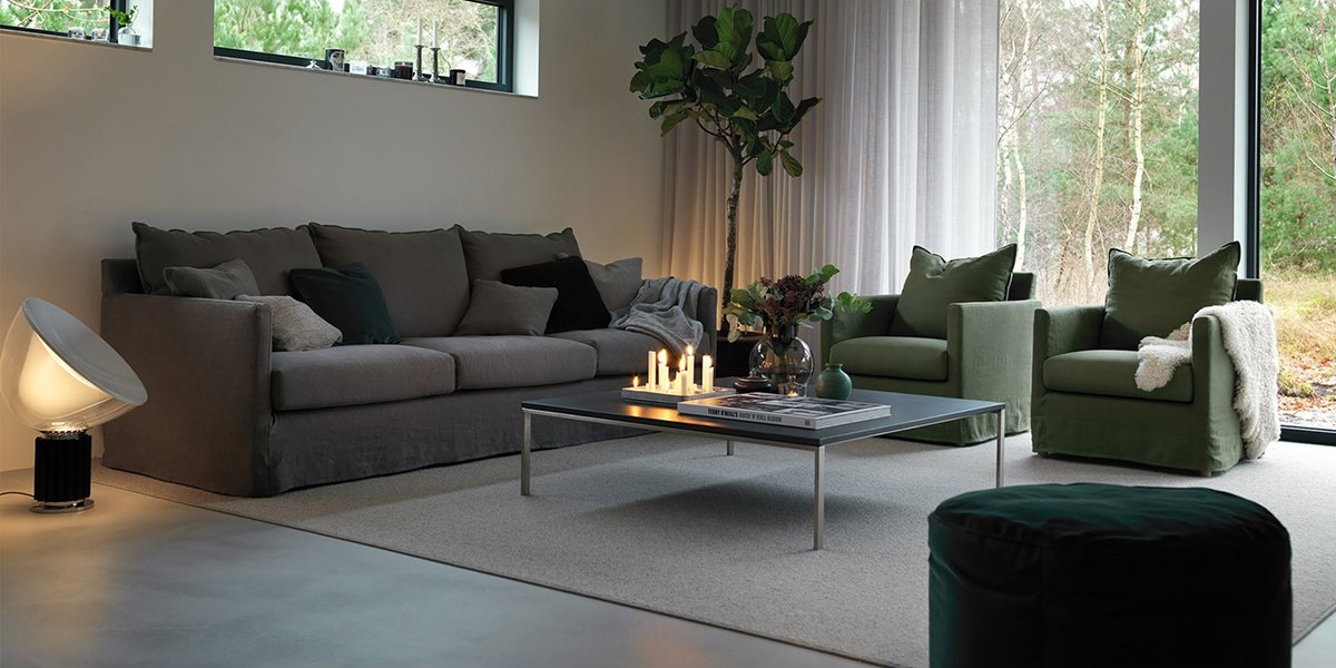 Salon moderne avec un canapé gris élégant, des fauteuils verts chics, des bougies allumées sur la table basse et une grande fenêtre offrant une vue rappelant une salle d'exposition parisienne avec des vues sereines sur la cime des arbres. Canape Paris by Atelier Bis