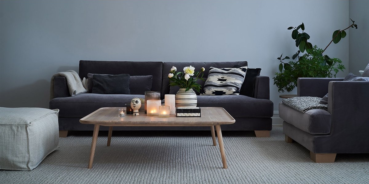 Un salon moderne d'inspiration parisienne comprend des canapés sombres, une table en bois ornée de bougies et de livres, et des plantes luxuriantes sur un tapis beige, dégageant un air de qualité tranquille. Canape Paris by Atelier Bis