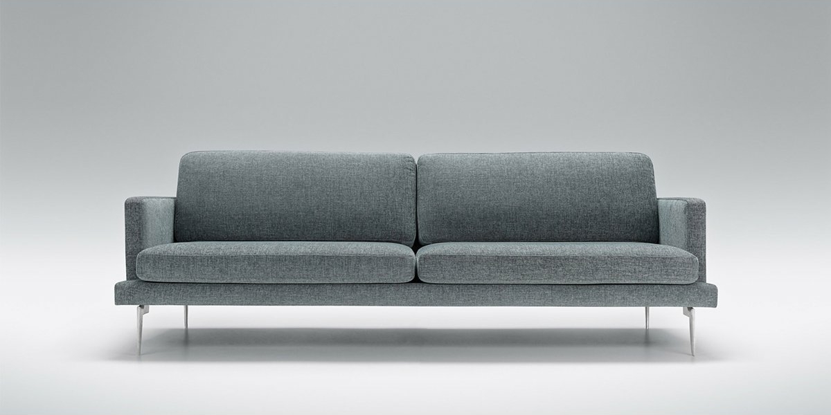 Un canapé moderne gris avec des pieds chromés se dresse élégamment sur un fond gris clair, offrant une touche de style parisien et une qualité inégalée. Canape Paris by Atelier Bis