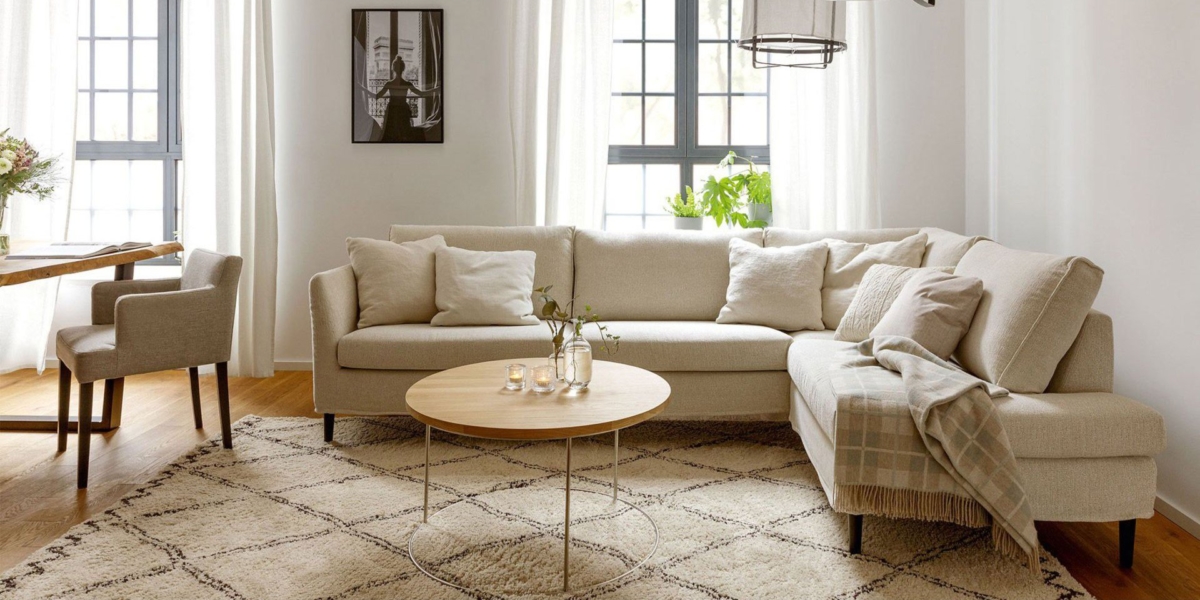 Salon confortable avec un canapé sectionnel beige de haute qualité, une table ronde en bois et un tapis à motifs dans un espace lumineux et vitré. Canape Paris by Atelier Bis