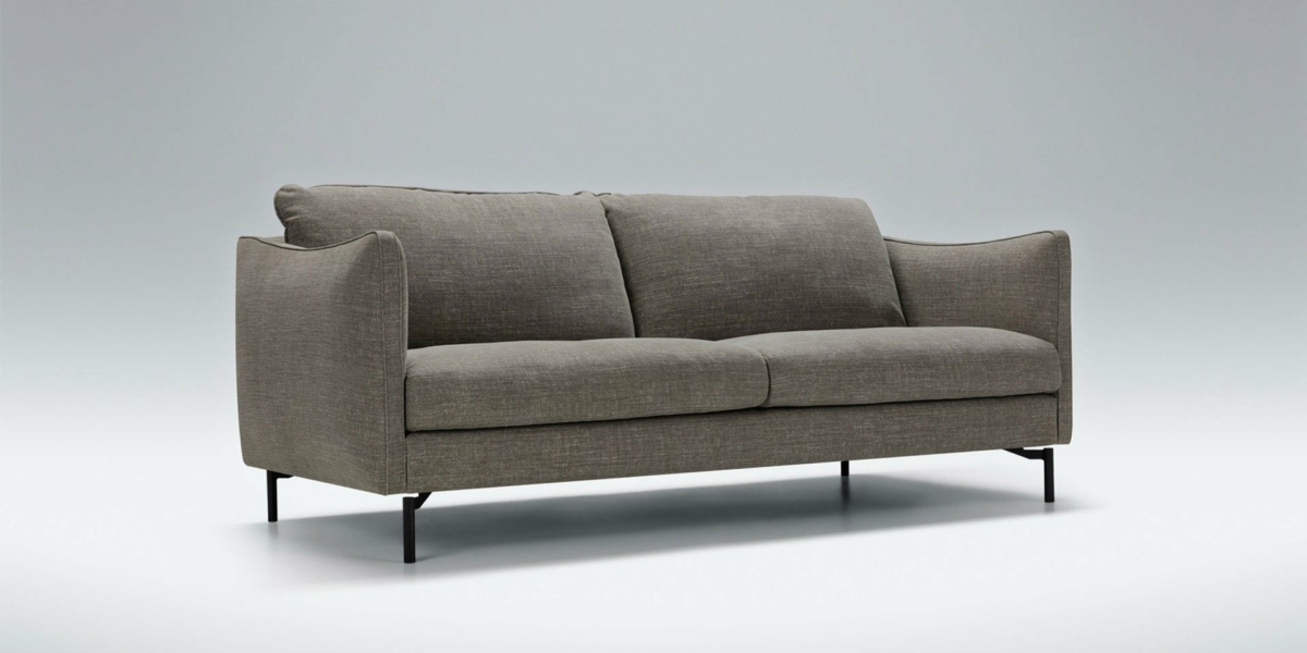Canapé gris moderne aux pieds noirs élancés, présenté dans une salle d'exposition sur un fond gris clair, dégageant un sentiment de qualité et d'élégance contemporaine. Canape Paris by Atelier Bis