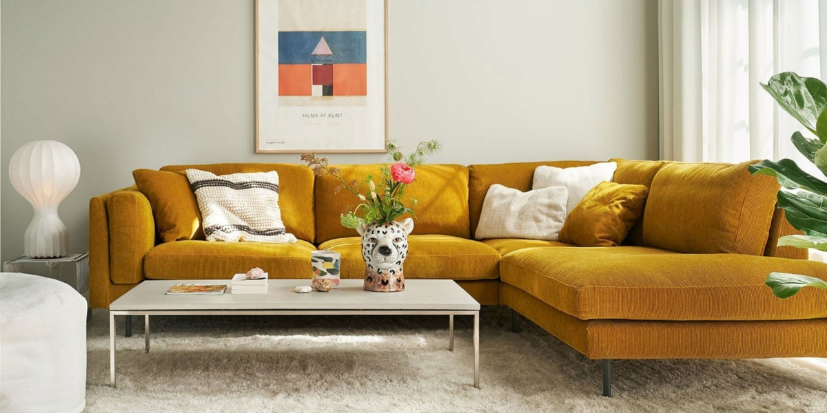 Le charme parisien orne ce salon avec un canapé sectionnel jaune moutarde, une table basse blanche, des œuvres d'art murales abstraites et une plante en pot, le tout reflétant l'élégance de qualité d'une salle d'exposition. Canape Paris by Atelier Bis