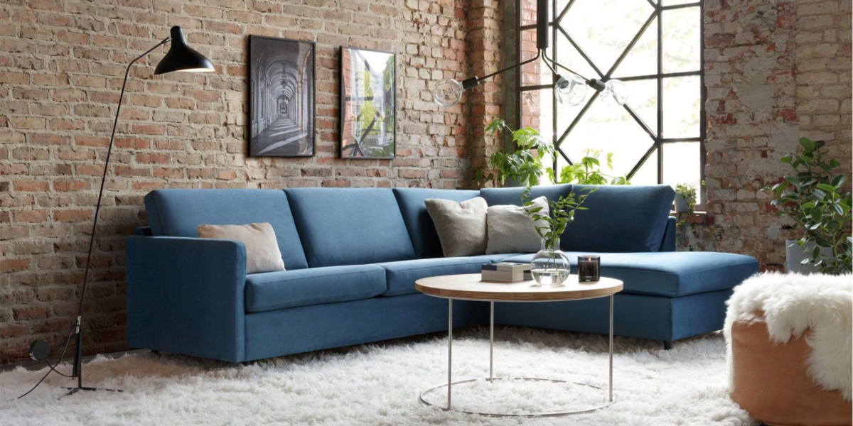 Salon cosy avec canapé bleu, murs en briques et table ronde en bois ornée de plantes. Un lampadaire moderne orne le tapis blanc moelleux, créant un charme digne d'un showroom rappelant l'élégance parisienne. Canape Paris by Atelier Bis