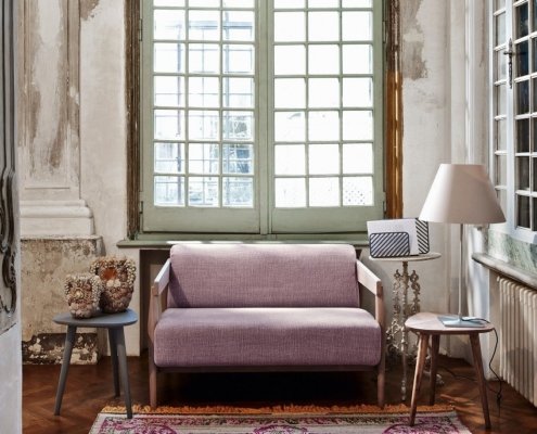 Une pièce cosy avec un canapé lavande, deux tables d'appoint, une lampe et un tapis coloré près de grandes fenêtres vintage respirant la qualité parisienne. Canape Paris by Atelier Bis