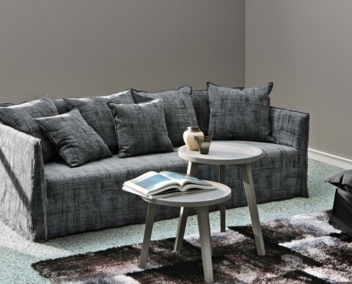 Canapé gris avec plusieurs coussins, deux tables d'appoint rondes blanches avec des livres et des décorations, et murs gris dans un showroom moderne rappelant l'élégance parisienne. Canape Paris by Atelier Bis