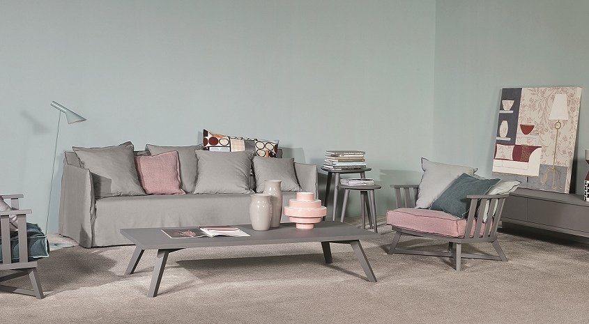 Un salon moderne présente des meubles gris, notamment un canapé et des fauteuils de haute qualité. Une table basse sophistiquée et des objets de décoration soigneusement choisis complètent les œuvres d'art qui ornent le mur gris, mélangeant ainsi les styles sans effort. Canape Paris by Atelier Bis