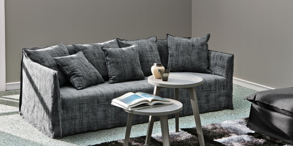 Canapé gris avec plusieurs coussins, deux tables d'appoint rondes blanches sur lesquelles reposent un livre et un vase, le tout élégamment disposé sur un tapis et une moquette texturés. Ce décor inspiré d'un showroom respire l'ambiance chic de la vie parisienne. Canape Paris by Atelier Bis