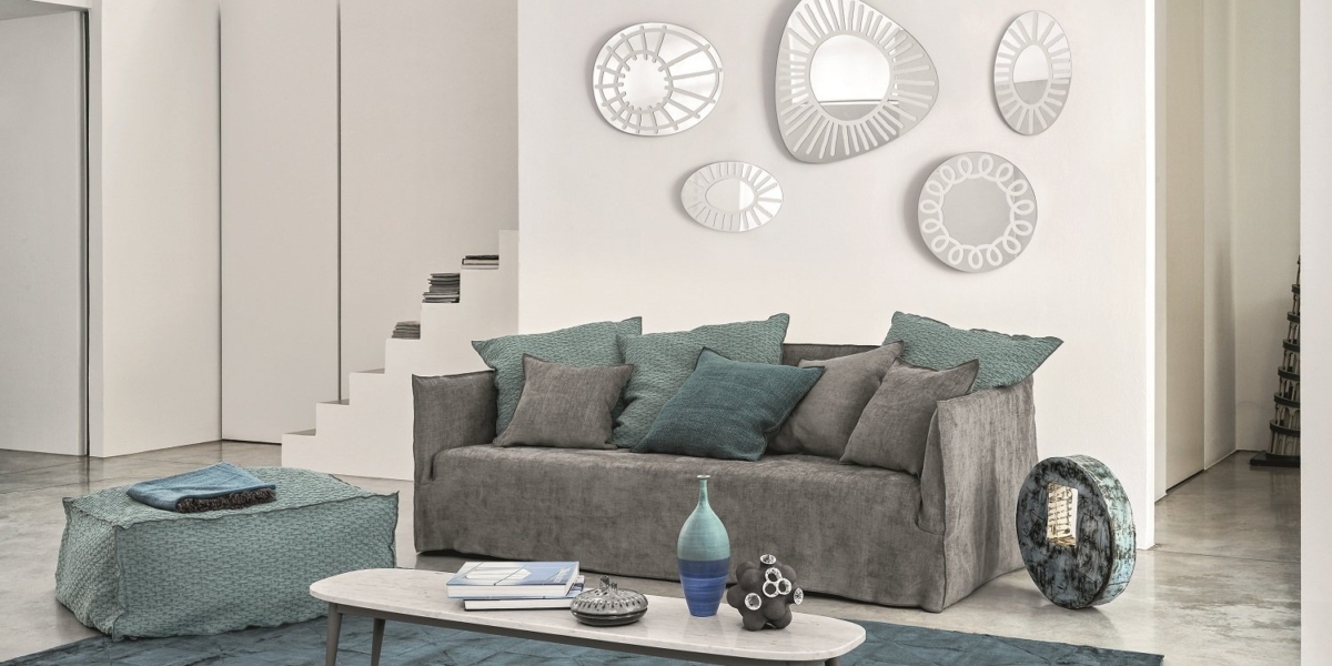 Un salon moderne comprend un canapé gris orné de coussins bleu sarcelle, des miroirs décoratifs, une table basse et un escalier sur un sol poli. Canape Paris by Atelier Bis