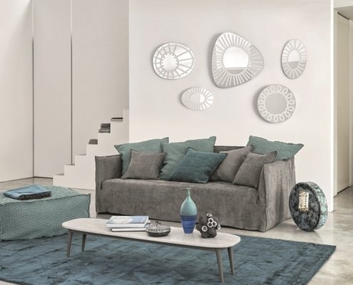 Découvrez un salon moderne doté d'un élégant canapé gris orné de coussins bleu sarcelle, de miroirs muraux abstraits et d'une table basse minimaliste, le tout faisant écho à la sophistication d'un showroom haut de gamme. Canape Paris by Atelier Bis