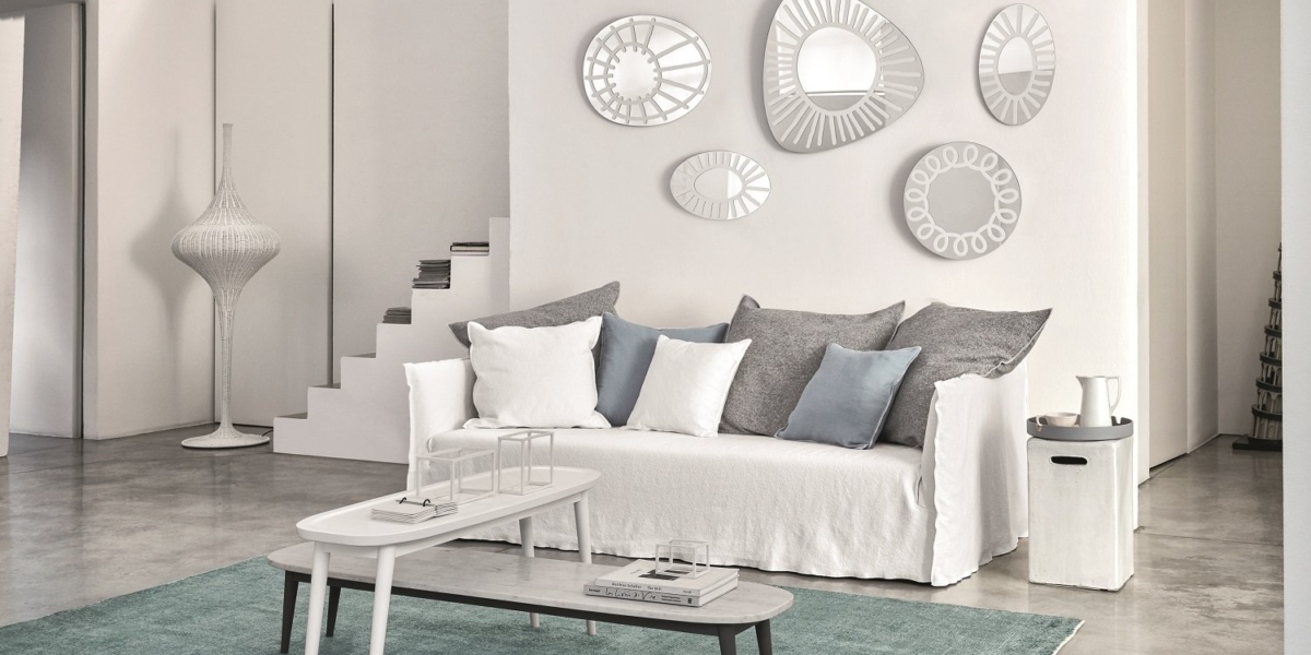 Salon minimaliste comprenant un canapé blanc, des coussins gris, des miroirs décoratifs au mur et des tables gigognes sur un tapis. Canape Paris by Atelier Bis