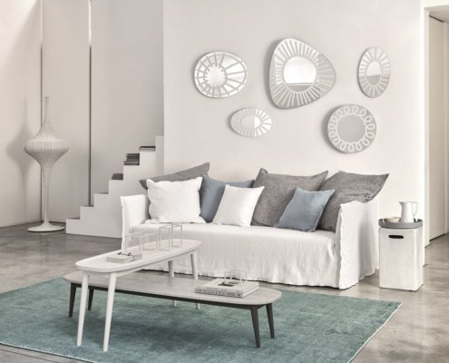 Salon minimaliste comprenant un canapé blanc, des coussins gris, des miroirs décoratifs au mur et des tables gigognes sur un tapis. Canape Paris by Atelier Bis