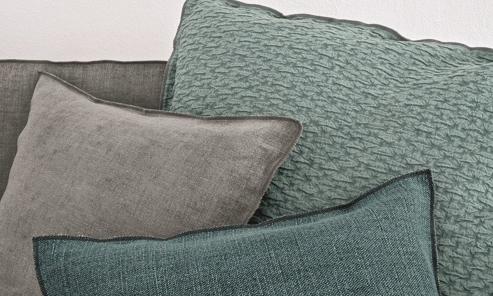 Un groupe de coussins texturés dans des tons gris et bleu sarcelle est élégamment disposé contre un mur blanc, dégageant une qualité semblable à celle d'une salle d'exposition. Canape Paris by Atelier Bis