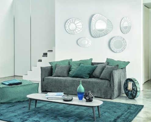 Salon moderne présentant la qualité d'une salle d'exposition, avec un canapé gris orné de coussins bleu sarcelle, une œuvre d'art murale abstraite, une table basse élégante et un tapis bleu sarcelle sur le sol en béton. Canape Paris by Atelier Bis