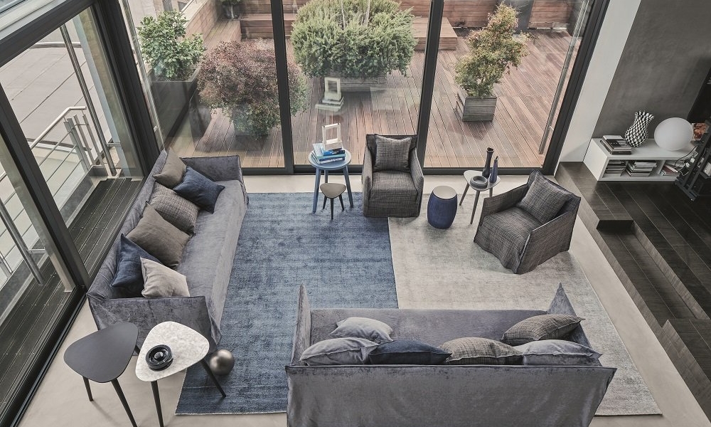 Ce salon moderne ressemble à un showroom parisien avec ses canapés gris, son tapis bleu et ses grandes fenêtres donnant sur un patio orné de plantes luxuriantes, offrant une ambiance d'une qualité inégalée. Canape Paris by Atelier Bis