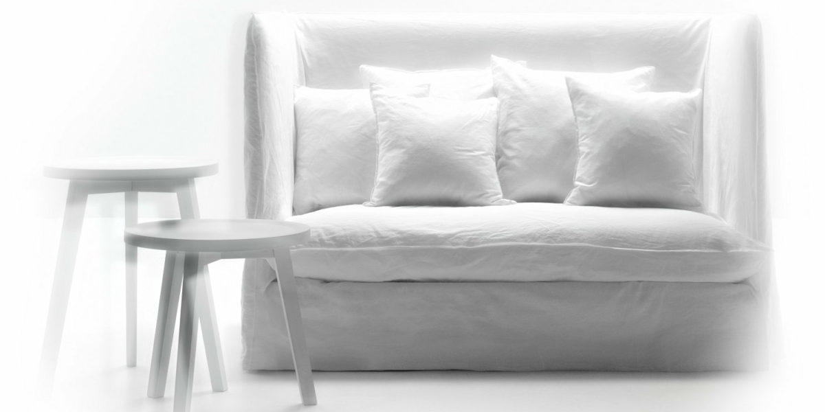 Dans un espace lumineux et aéré, un canapé blanc minimaliste est orné de quatre coussins et flanqué de deux petites tables rondes blanches, évoquant l'élégance sophistiquée d'un showroom parisien. Canape Paris by Atelier Bis