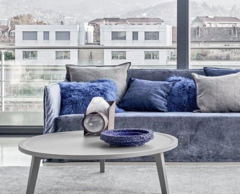 Découvrez un salon parisien moderne avec une table basse grise chic, des objets de décoration élégants et un canapé bleu moelleux. De grandes fenêtres offrent une vue imprenable sur le paysage urbain, alliant sophistication et charme chaleureux. Canape Paris by Atelier Bis