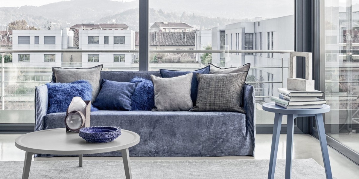 Un salon moderne à Paris comprenant un canapé bleu de qualité, divers coussins, deux tables rondes et de grandes fenêtres offrant une vue imprenable sur le paysage urbain. Canape Paris by Atelier Bis