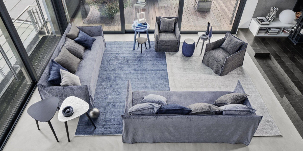 Un salon parisien chic et moderne présente deux canapés bleus, appelés en français « canapés », accompagnés de deux fauteuils et d'une table basse sur un tapis bleu et gris, offrant une vue élégante sur le patio. Canape Paris by Atelier Bis