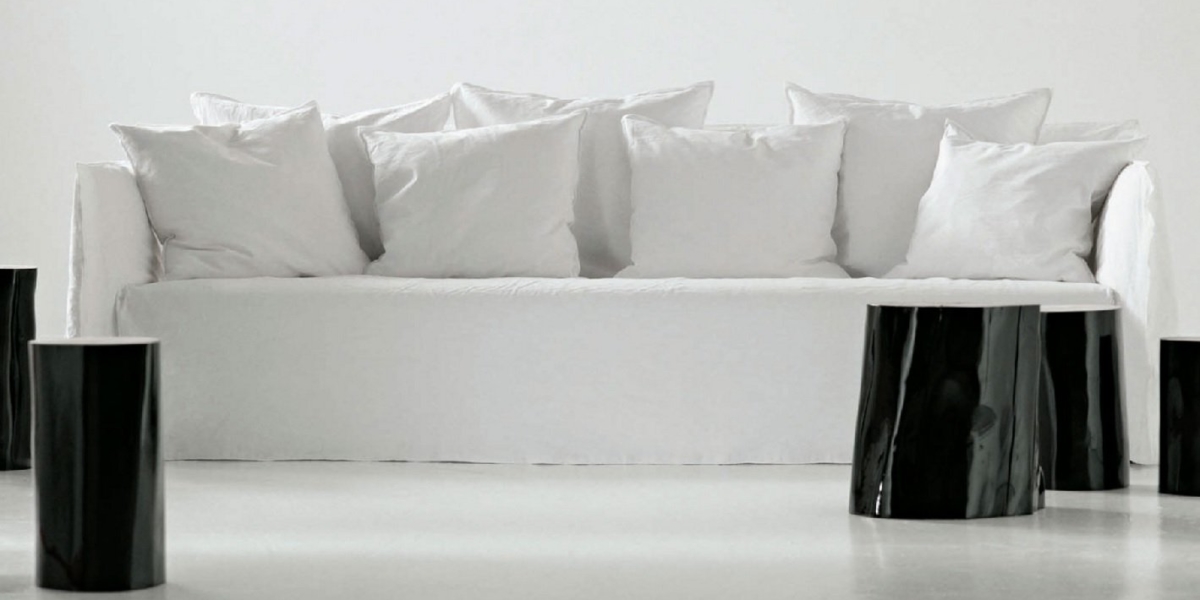 Canapé blanc avec plusieurs coussins blancs, entouré de tables d'appoint cylindriques noires épurées dans un cadre minimaliste qui respire la qualité chic du design parisien. Canape Paris by Atelier Bis