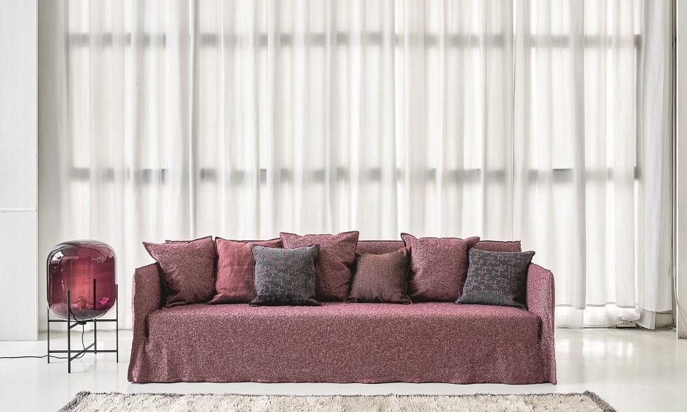 Un canapé marron orné de coussins assortis trône gracieusement devant des rideaux transparents, à côté d'une table d'appoint ornée d'un dôme en verre chic, parfait pour tout décor de salle d'exposition élégant. Canape Paris by Atelier Bis