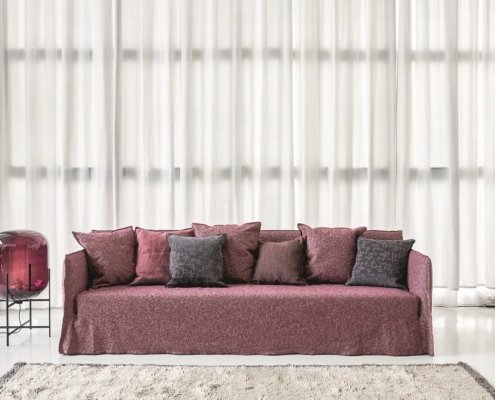 Un canapé marron orné de coussins assortis trône gracieusement devant des rideaux transparents, à côté d'une table d'appoint ornée d'un dôme en verre chic, parfait pour tout décor de salle d'exposition élégant. Canape Paris by Atelier Bis