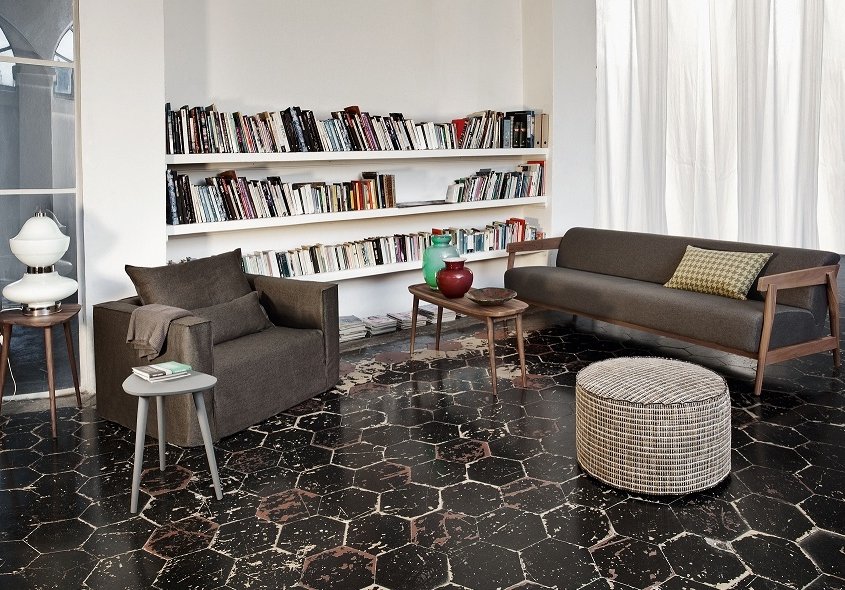 Salon cosy rappelant un showroom parisien, comprenant des étagères, un canapé gris, un fauteuil, un pouf rond et une table d'appoint sur un sol à motifs. Canape Paris by Atelier Bis