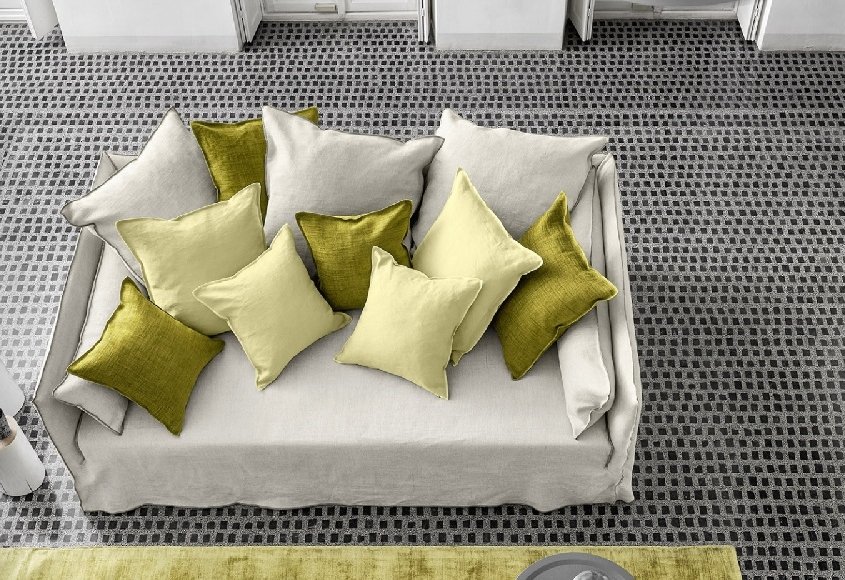 Retrouvez la qualité du design parisien avec un canapé beige agrémenté de coussins verts et jaunes sur un sol à carreaux. Ce décor élégant est complété par des tabourets cylindriques blancs, créant une ambiance digne d'un showroom. Canape Paris by Atelier Bis