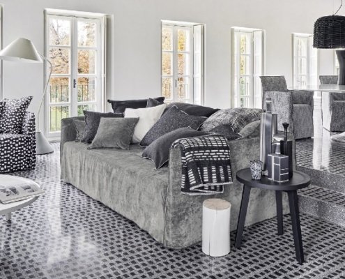 Salon moderne avec mobilier gris et coussins à motifs, rappelant un showroom parisien. Le sol carrelé complète un canapé confortable, tandis que le coin repas est élégamment installé sur une plate-forme surélevée. Canape Paris by Atelier Bis