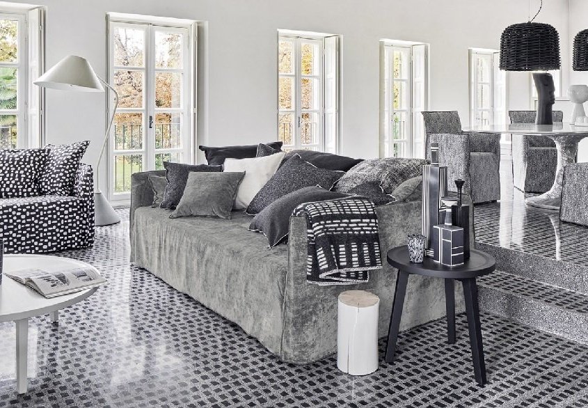 Salon moderne avec mobilier gris et coussins à motifs, rappelant un showroom parisien. Le sol carrelé complète un canapé confortable, tandis que le coin repas est élégamment installé sur une plate-forme surélevée. Canape Paris by Atelier Bis