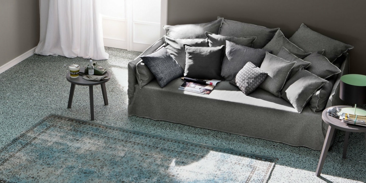 Un salon confortable avec un canapé gris rempli d'oreillers, deux petites tables rondes, un tapis à motifs et une fenêtre avec des rideaux dignes de n'importe quelle salle d'exposition. Canape Paris by Atelier Bis