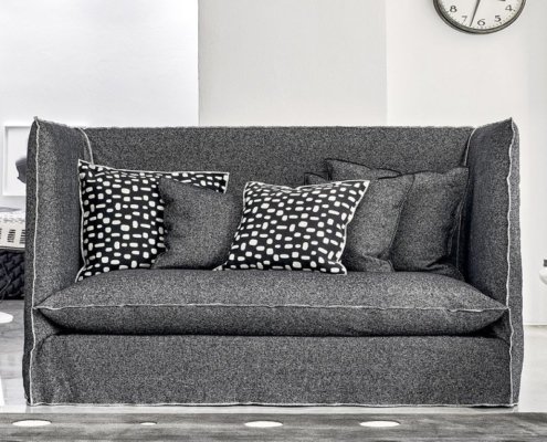 Un élégant canapé gris, orné de coussins à motifs noirs et blancs, orne cette pièce moderne rappelant un showroom parisien, avec une élégante horloge murale. Canape Paris by Atelier Bis