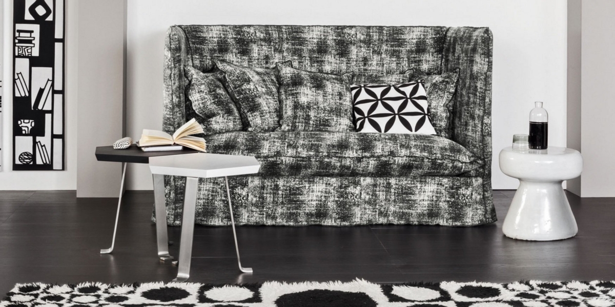 Canapé gris texturé avec coussins, motifs géométriques en noir et blanc et livre ouvert. Table et tabouret d'appoint à proximité, rappelant un appartement parisien chic. Canape Paris by Atelier Bis